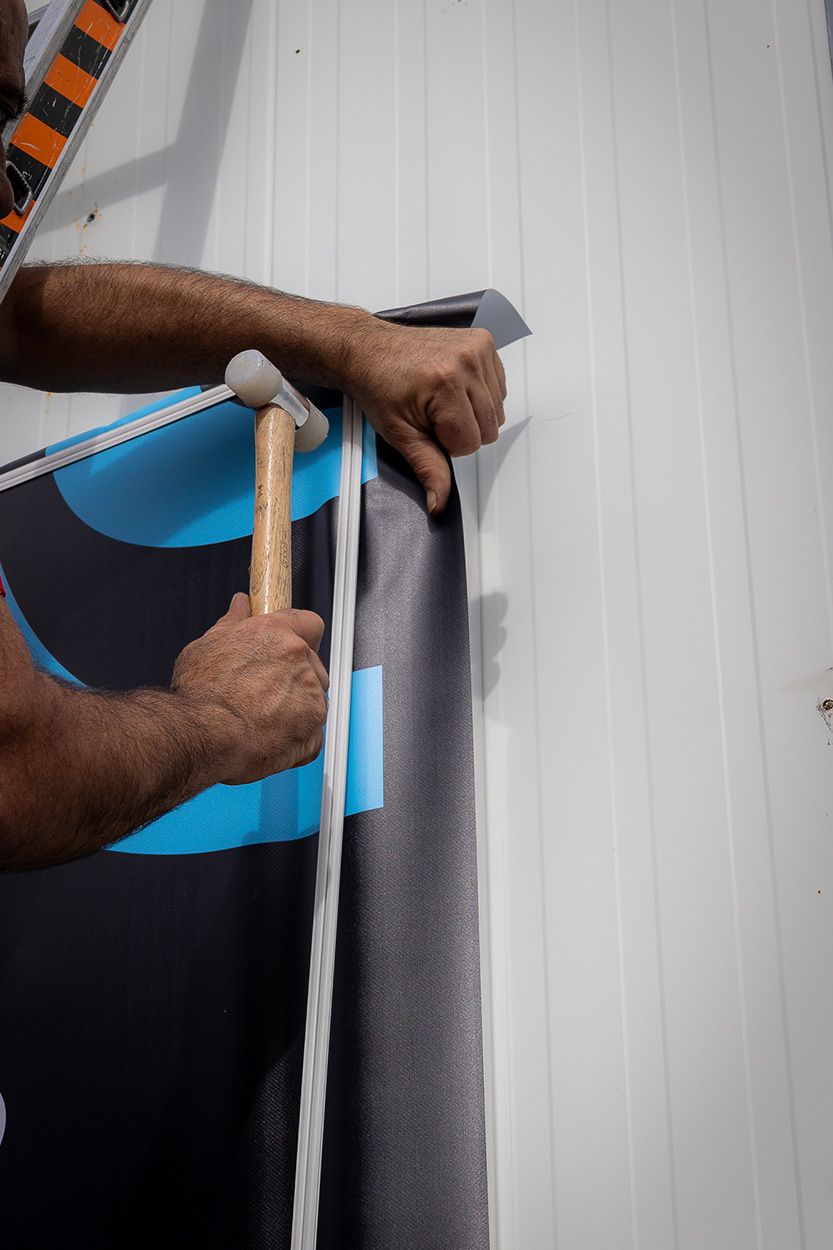 Artisan poseur installant une toile tendue imprimée sur un profilé mural à l'aide d'un maillet, fabrication et pose de supports de communication vers Mont-de-Marsan par Griffe Communication