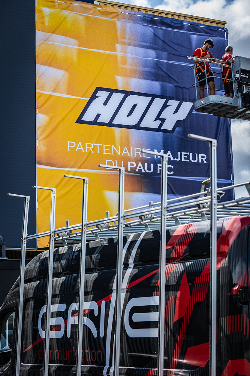 Équipe de poseurs sur nacelle installant une bâche géante en toile tendue pour la marque Holy au Pau FC avec le fourgon Griffe Communication au premier plan, création de signalétique extérieure à Pau